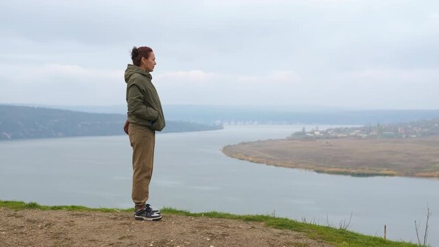 Woman enjoying the serene landscape from a high viewpoint. Thoughtful woman standing on a windy cliff edge, gazing at a vast river bend and distant landscape on a cloudy, atmospheric day