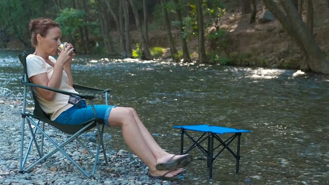 Enjoy a drink by the river in summer. Mature woman resting by gently flowing river, seated in comfortable camping chair, holding warm beverage and experiencing peaceful outdoors landscape