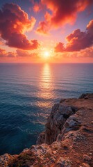 Fototapeta premium Dramatic sunset over calm ocean viewed from rugged cliff edge. Vibrant orange clouds fill sky above sea water. Golden sunlight reflects on waves during evening. Scenic coastal landscape with rocky