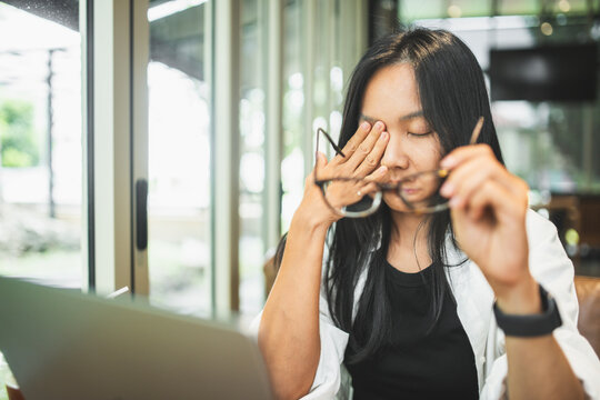 Tired woman removing glasses and rubbing her eyes while working on a laptop, illustrating eye strain, visual fatigue, prolonged screen time, stress and digital work.