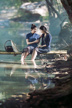 Two happy Asian women in traditional indigo dress catching fish on a bamboo bridge in Sakon Nakhon, Thailand. Perfect for travel banners and culture ads.