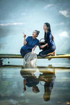 Two happy Asian women in traditional indigo dress catching fish on a bamboo bridge in Sakon Nakhon, Thailand. Perfect for travel banners and culture ads.