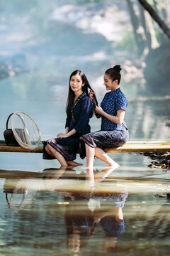 Two happy Asian women in traditional indigo dress catching fish on a bamboo bridge in Sakon Nakhon, Thailand. Perfect for travel banners and culture ads.