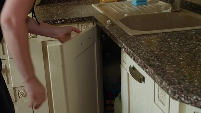 Person throwing garbage into waste container beneath kitchen sink counter with granite countertop, soap dispenser, dish rack visible in residential kitchen setting.