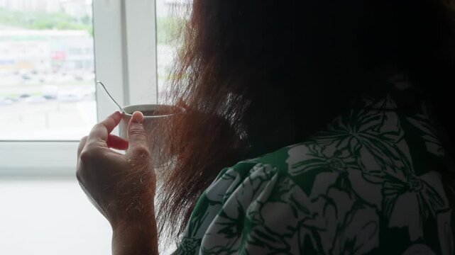Woman in green patterned clothing holds ceramic cup by large window overlooking urban landscape, captured from behind during quiet morning moment.