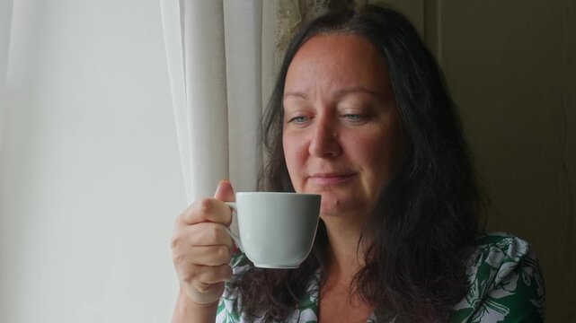 Adult woman sips hot beverage from white ceramic mug indoors near window during morning, peaceful quiet moment at home.