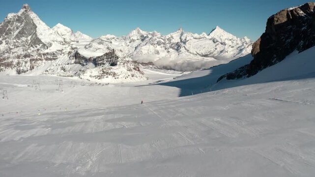 Skier descends an empty glacier slope in Zermatt with the iconic Matterhorn rising above spectacular snow-covered peaks under clear blue skies in the Swiss Alps.