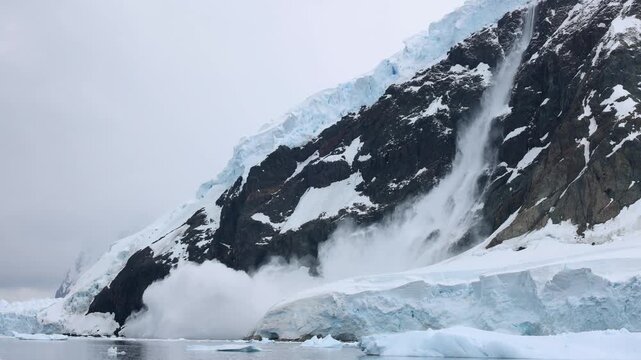 Snow and ice cascade dramatically down a rugged, black-rock cliff in Antarctica, creating a powerful avalanche. Overcast skies, frigid landscape, and cinematic composition enhance the spectacle.