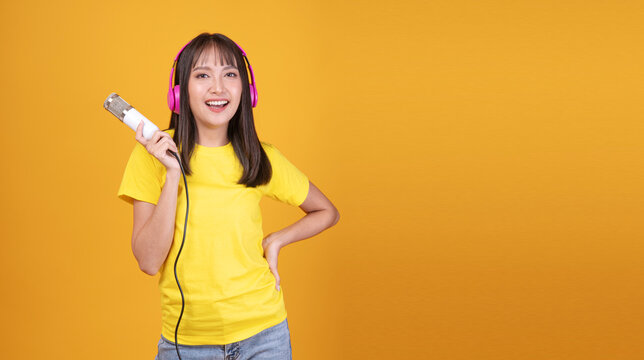 Joyful young Asian woman wearing headphones holding microphone singing isolated on yellow background with copy space for podcast, audio or music banner.