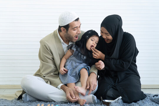 Happy Muslim family sitting together on carpet, mother feeding little daughter while father watching cheerful, isolated on white room with copy space.