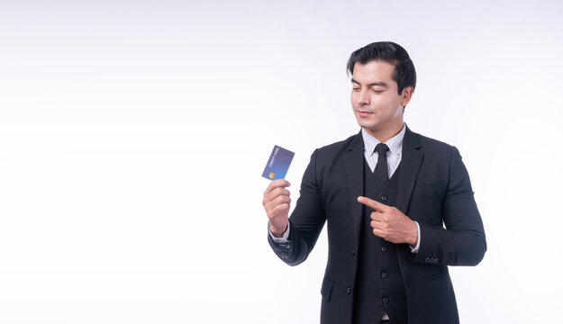 Confident businessman in elegant black suit pointing finger at credit card isolated on white background with negative copy space for financial advertising.