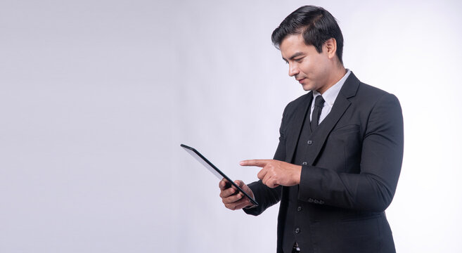 Focused businessman in black suit holding digital tablet and pointing finger at screen isolated on white background with copy space for technology corporate app.