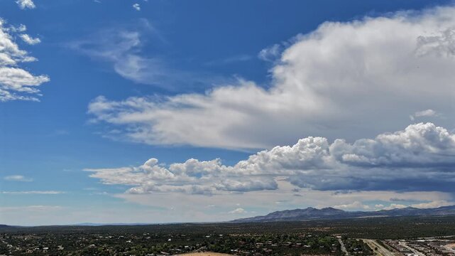 A 4k drone timelapse capturing multiple cloud layers shifting dynamically in a bright blue sky over a sprawling New Mexico town and a distant mountain range.