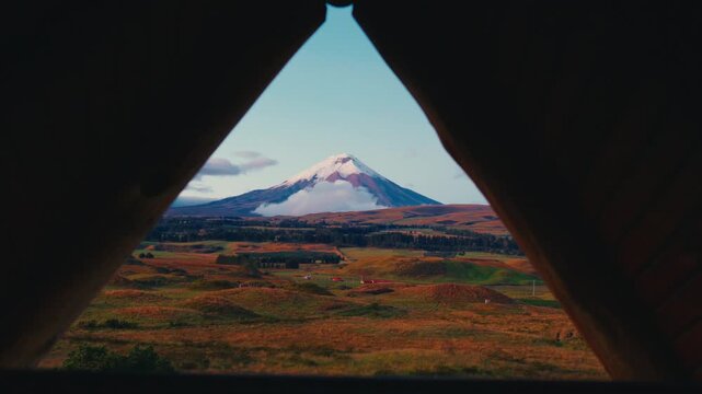 Cotopaxi volcano viewed through a triangular wooden frame with rolling hills and clouds below