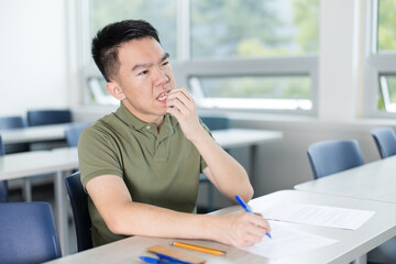 A young Asian male college student bites his nails while completing a written test in a classroom, for academic pressure, exam preparation, student anxiety, or education concepts. © fotolite
