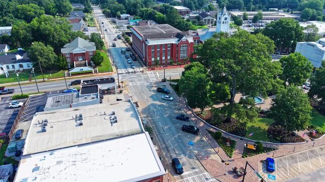 Cars travel busy street past red brick storefronts toward landscaped town square in Atlanta, Georgia, courthouse building and church steeple rise above flat rooftops. Aerial drone flyover shot
