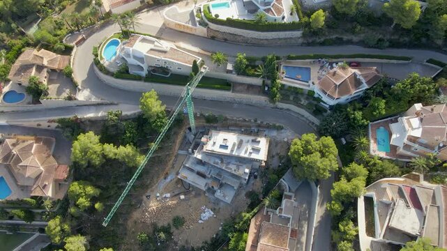Drone view showing a luxury villa under construction with a crane, surrounded by other houses with pools and winding roads.