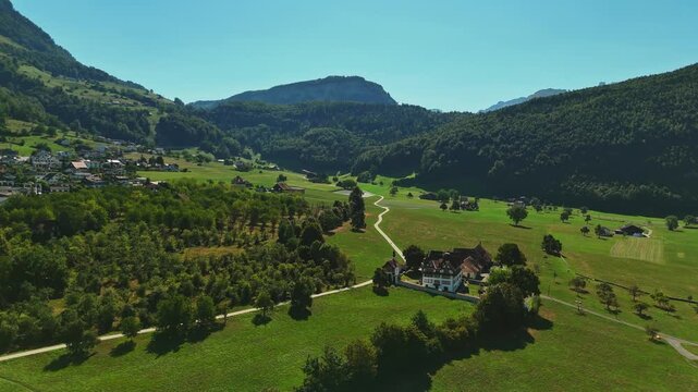 Aerial view of green countryside near Schwyz, Switzerland, with orchards, scattered farmhouses, winding rural lanes and forested Alpine hills on a clear summer day.