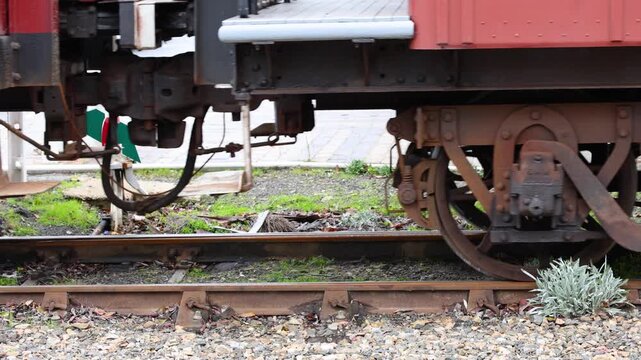 Detailed close-up shot of an old train wagon&rsquo;s wheels and undercarriage moving along rusty tracks, with visible springs, metal textures, and gravel under natural daylight.