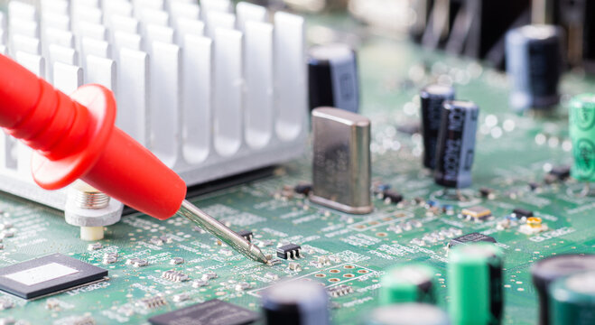 Red multimeter probe measuring a tiny electronic component on a green printed circuit board