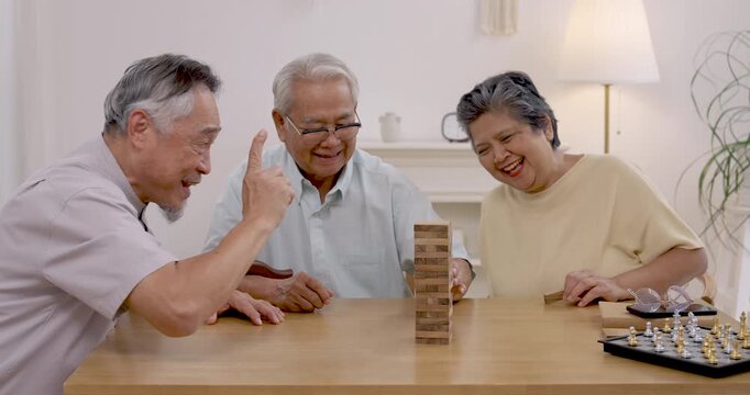 Asian senior friends laugh and take turns moving wooden blocks during social recreation in modern assisted living center, enjoying active aging, friendship, playful interaction and companionship.