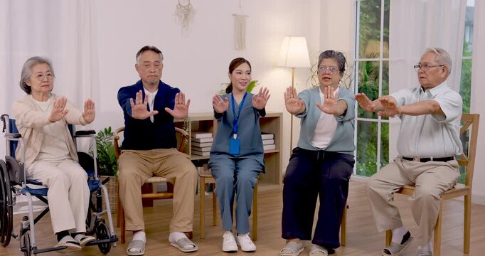 Senior Asian group performs seated arm exercise with nurse guidance, moving arms forward and upward during gentle rehabilitation activity inside care center supporting mobility and healthy aging.