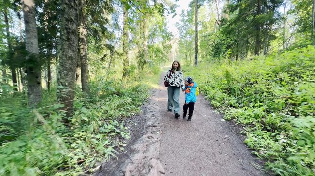 A mother and young son walk along a dirt path at Valaam near Lake Ladoga. Tall green pine trees surround them in the quiet woods. Their peaceful outdoor stroll brings warm family joy and happiness.