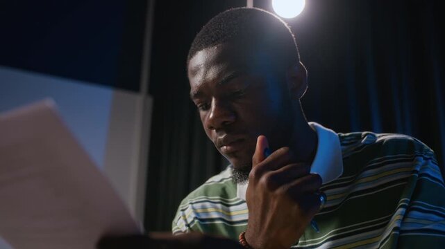 African American young adult student holding pen while reading theater script during stage rehearsal, zoom out. Suitable for drama school advertising and arts education promo campaigns