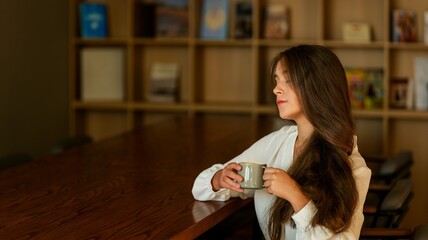 Young woman with long brown hair holding a coffee mug while sitting at a wooden table © shine.graphics