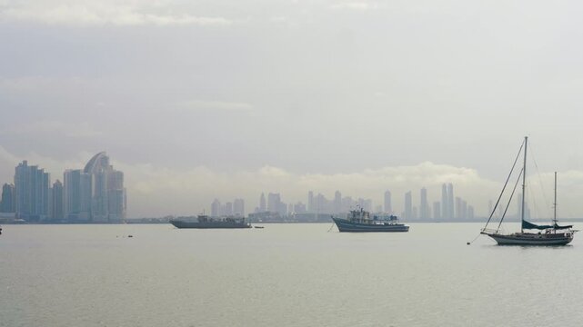 Calm waters with sailboats and cargo boats in the foreground; In the background, an imposing silhouette of skyscrapers wrapped in soft mist under a cloudy sky