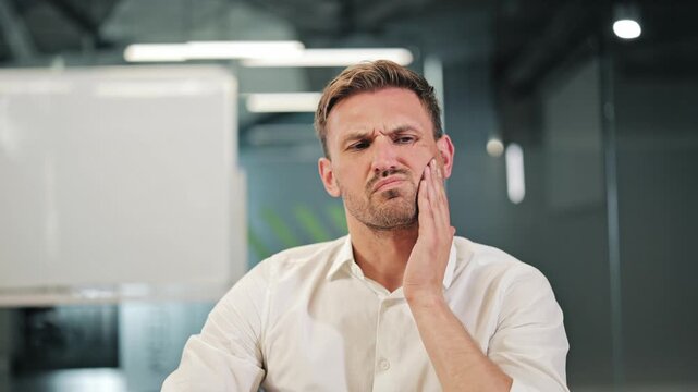Adult experiences intense dental pain while at work. Discomfort evident on face. Medical issue affecting productivity in workplace. Need for treatment apparent. Health concern disrupts daily routine.