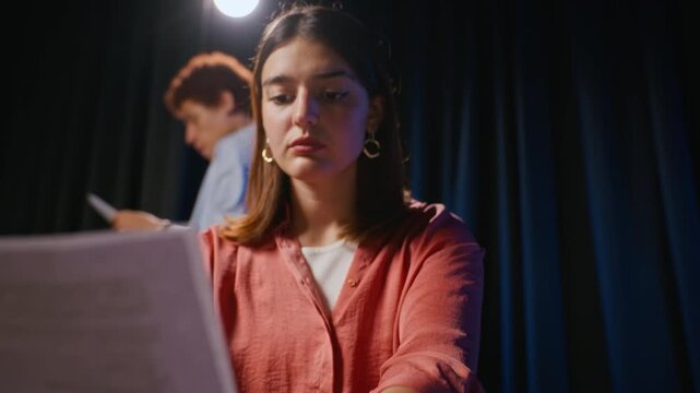 Caucasian young adult woman reading script pages during theater rehearsal while actor practices lines in background. Footage fits drama school advertising or performing arts education campaigns