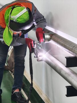 A medium shot of an industrial worker bending over to perform precise TIG welding on a wall-mounted stainless steel pipe line, producing a bright arc glare while wearing a flip-up face shield