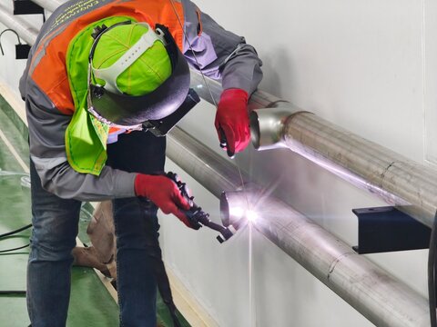 A medium shot of an industrial worker bending over to perform precise TIG welding on a wall-mounted stainless steel pipe line, producing a bright arc glare while wearing a flip-up face shield