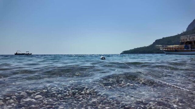 Clear sea waves roll over a pebble shore with a buoy, boats and a ship in the distance, captured in slow motion
