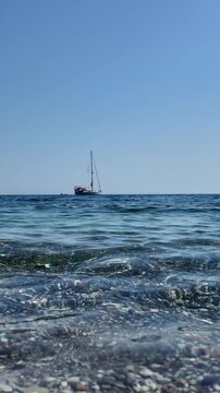 Sailboat on the horizon over clear blue sea with gentle waves, captured in vertical slow motion