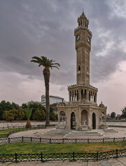 Fototapeta premium Historic Izmir Clock Tower at Konak Square in Izmir, Turkey, an iconic Ottoman landmark featuring intricate stone carvings and a prominent palm tree under a dramatic cloudy sky