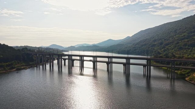 A bridge connects two land areas over a river. The sun shines and reflects off the water. Mountains rise in the background and the sky has clouds.