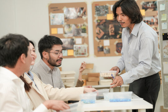 The seated men lean in as he points at the sample stack, in a daylit creative studio, an Asian architecture and design team collaborating on an environmental project.