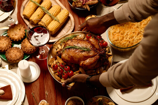 Hands of African American man serving roasted turkey at Thanksgiving dinner table. Image represents holiday catering, family gathering, and festive food marketing, high angle view
