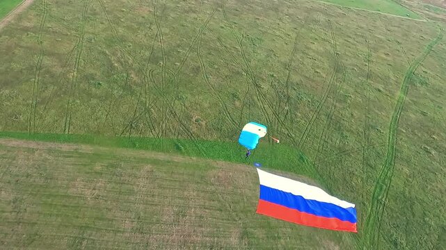 A bird&rsquo;s‑eye view of a parachutist under a canopy, with a large Russian tricolour, as they descend over green fields.
