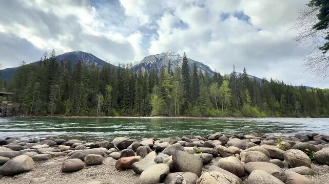 Static shot of a serene river flowing past a rocky shore with a dense forest and snow-capped mountains in the background under a cloudy sky, captured in Mount Robson Provincial Park, Canada.