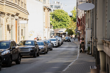 Historic townhouses frame a narrow Riga street where a single flag hangs overhead, parked cars and passers-by giving the scene its ordinary, lived-in character. © K