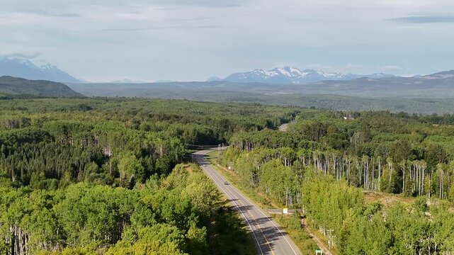Aerial shot of a two-lane road cutting through a dense forest with snow-capped mountains in the distance, showcasing clear, balanced framing and natural lighting in New Hazelton.