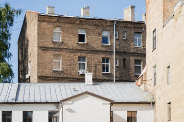 An old brick apartment building rises behind a tin rooftop, its weathered chimneys and rows of windows showing the patchwork repairs of a century-old structure. © K