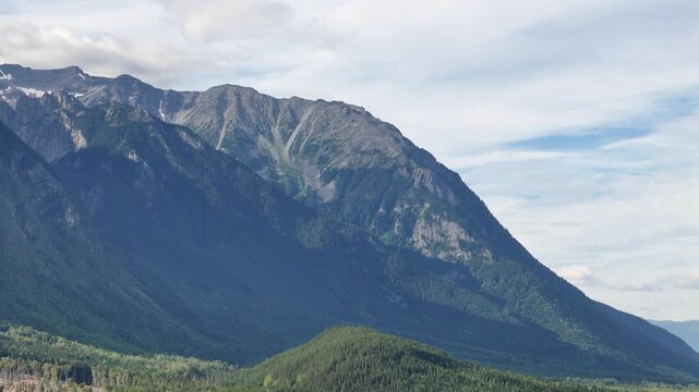 Aerial shot of a majestic mountain range covered in dense green forests at New Hazelton, under partly cloudy skies, evoking tranquility and vastness.