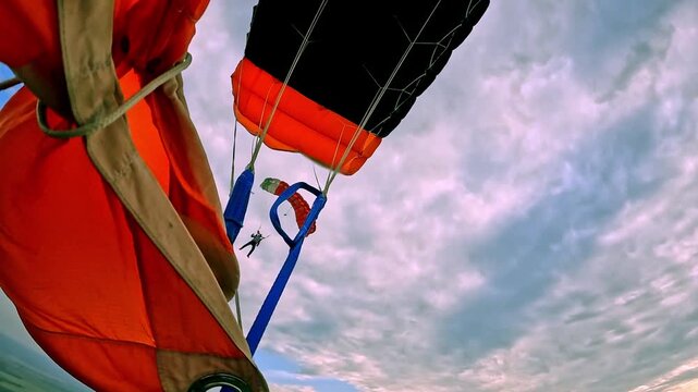 A view from under the canopy of another parachutist descending over the fields against the backdrop of a cloudy sky and the evening light.
