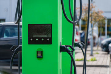 A green electric vehicle charging station stands curbside with its cables coiled and screen displaying status, a parked car waiting nearby to plug in. © K