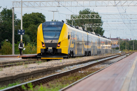 Commuter rail schedules hinge on moments like this one, a yellow regional train rolling toward the platform as a lone passenger waits near the signal post.