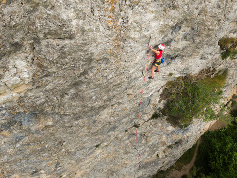 Climber ascends steep rock face. Red helmet stands out against gray stone. Moss patches cling to rock surface. Distant green valley below. Shows adventure spirit. Ideal for outdoor activity concepts